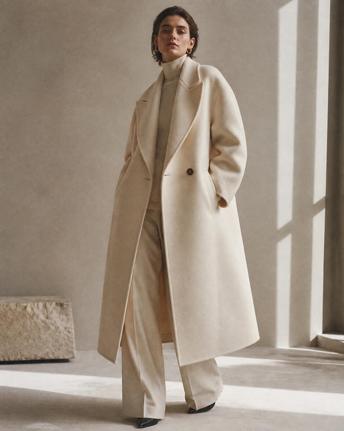 Model wearing an ivory double-breasted wool coat in a sunlit minimalist studio